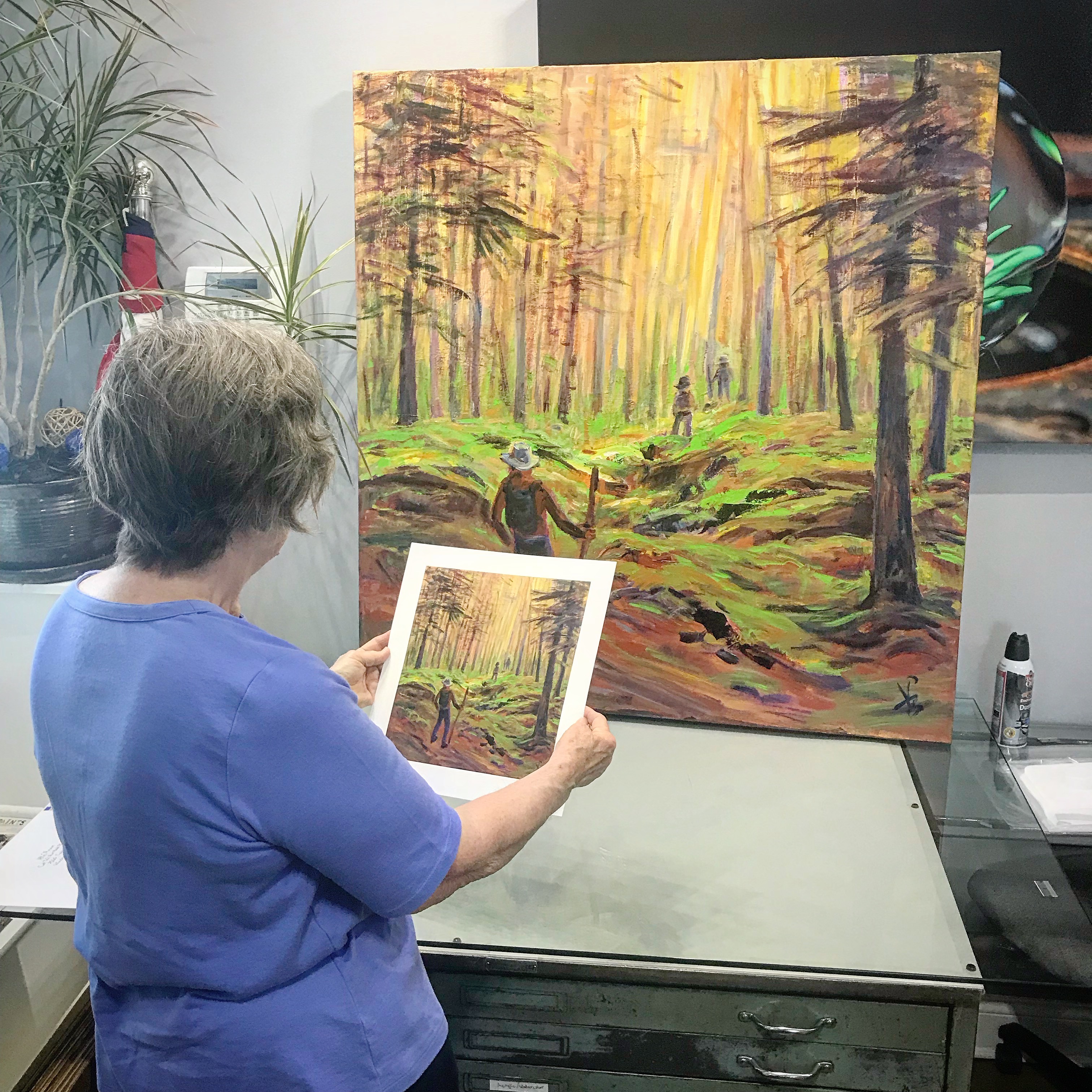 Artist Peg Bruhn reviewing a print proof next to her original artwork of hikers in a forest.